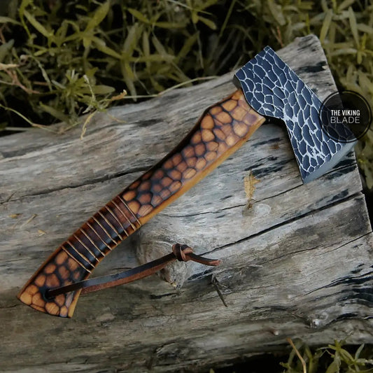 A small forged carbon steel axe with a burn ash wood shaft, displayed on a natural outdoor background.
