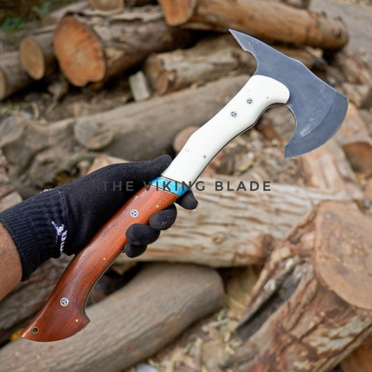 Hand holding a dual-bladed axe with wooden and white handles against a background of logs.