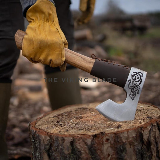 Hand holding a axe with decorative blade on a wooden stump, with blurred background of logs and nature.