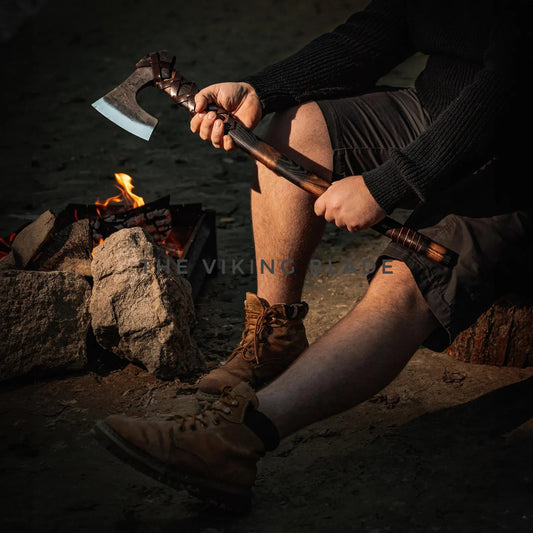 Person sitting by a campfire holding an axe, wearing boots and shorts.