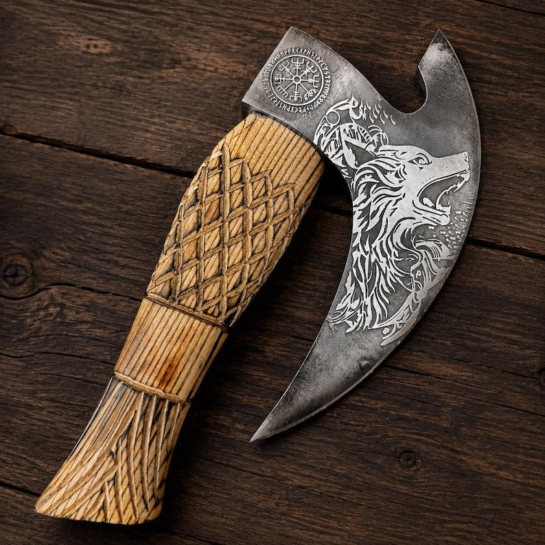 A Viking-style bearded axe with a carved wooden handle and a steel head featuring an engraved wolf and compass rose, displayed diagonally on a dark rustic wooden background.