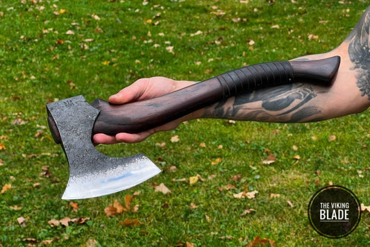 A person holding a Viking forged axe 'Asgard' with a brown leather handle and a steel head, outdoors on a grassy area.