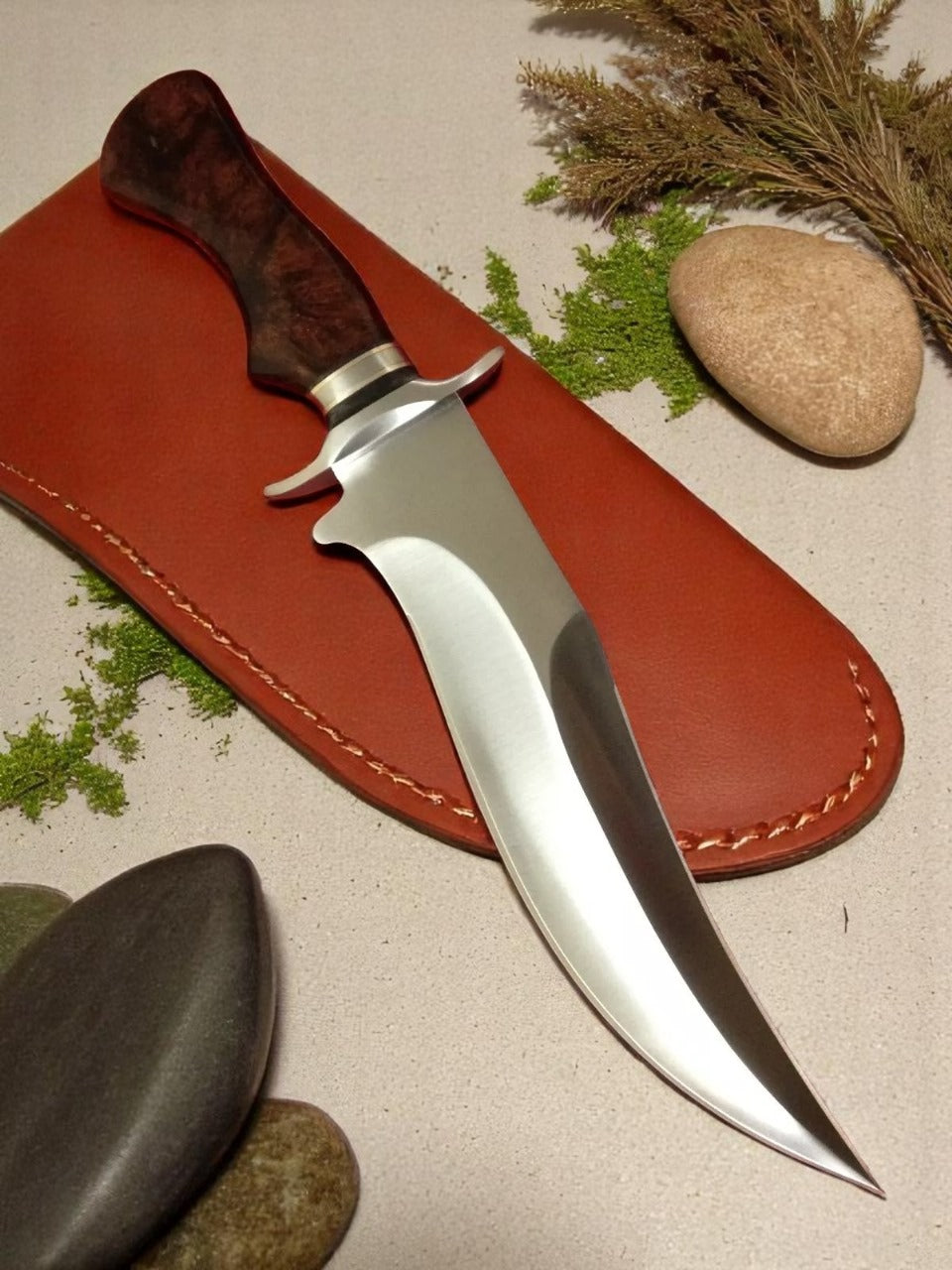 A custom handmade hunting Bowie knife with a silver blade and brown resin handle, accompanied by a brown leather sheath and displayed with stones and a plant in the background.
