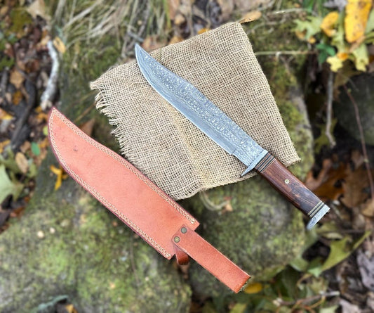 A custom handmade Damascus steel Bowie knife with a brown leather sheath, placed on a burlap fabric atop a rock.