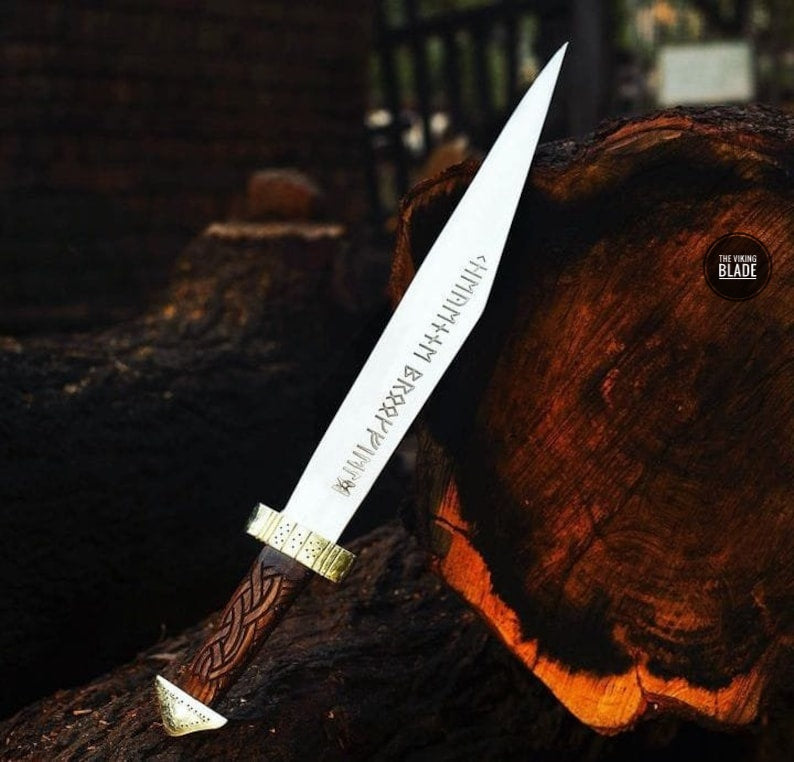 A hand-made Viking's Seax knife with a high carbon steel blade, rose wood handle, and brass pommel and guard, displayed on a wooden log.