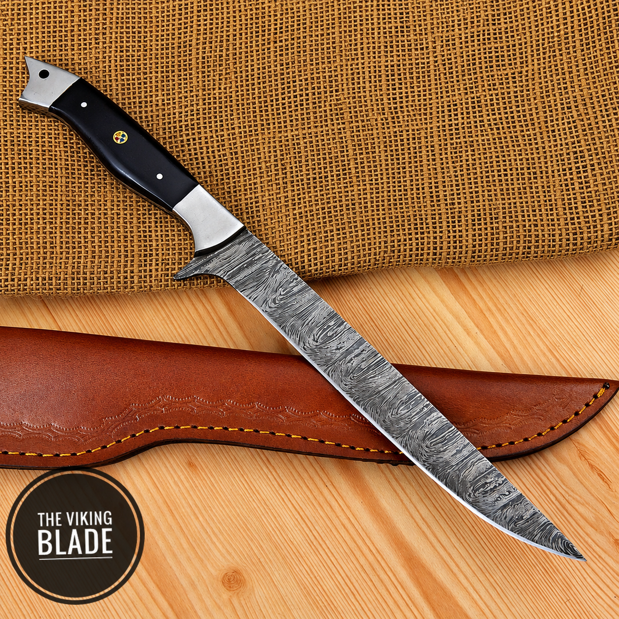 A fixed-blade knife with a patterned steel blade and a black handle with silver bolsters, displayed diagonally across a brown leather sheath on a light wood surface with a burlap cloth background. A circular logo in the bottom-left corner reads 'THE VIKING BLADE'.