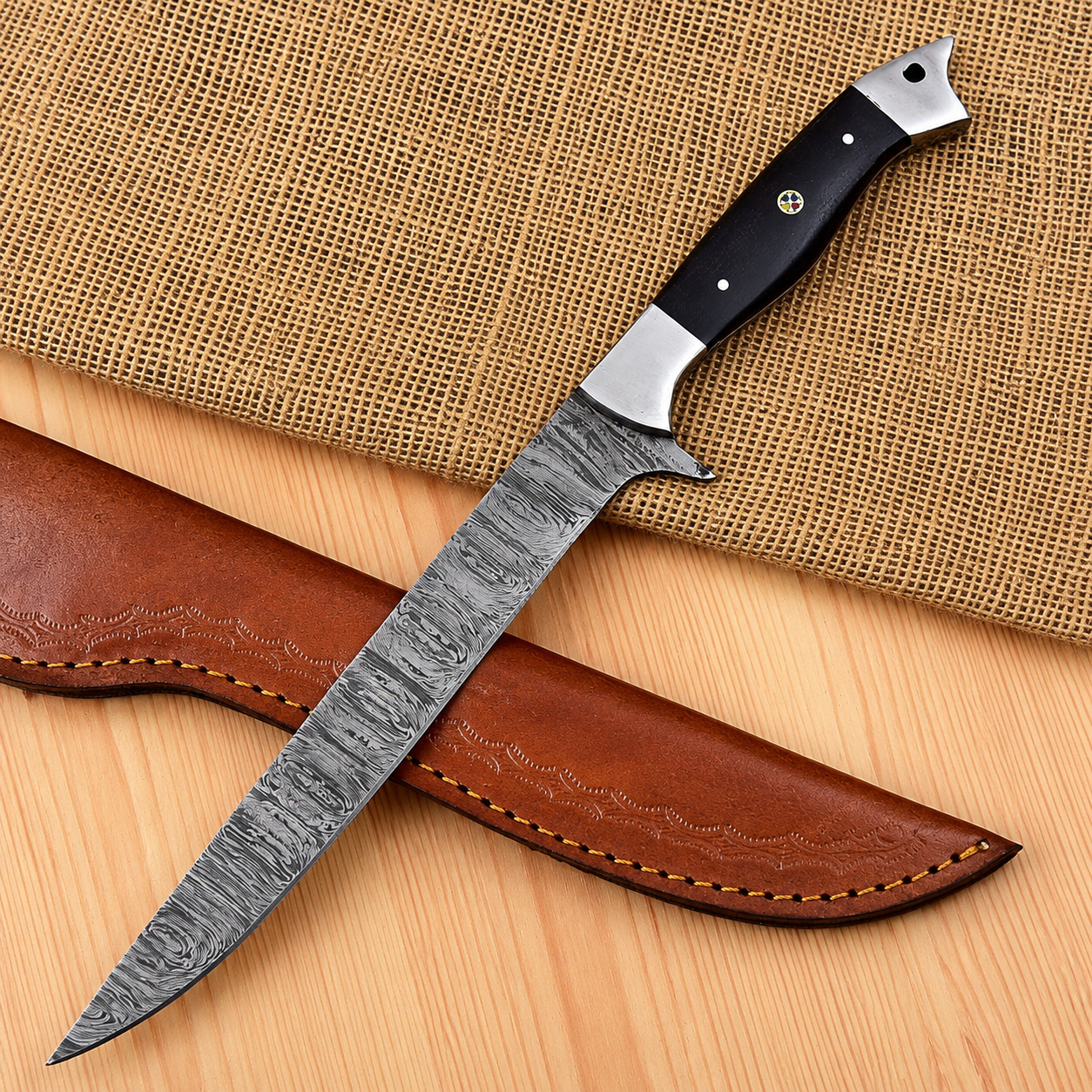 A fixed-blade knife with a patterned steel blade, a black handle with silver bolsters, shown resting on a brown leather sheath over a burlap and wood surface.