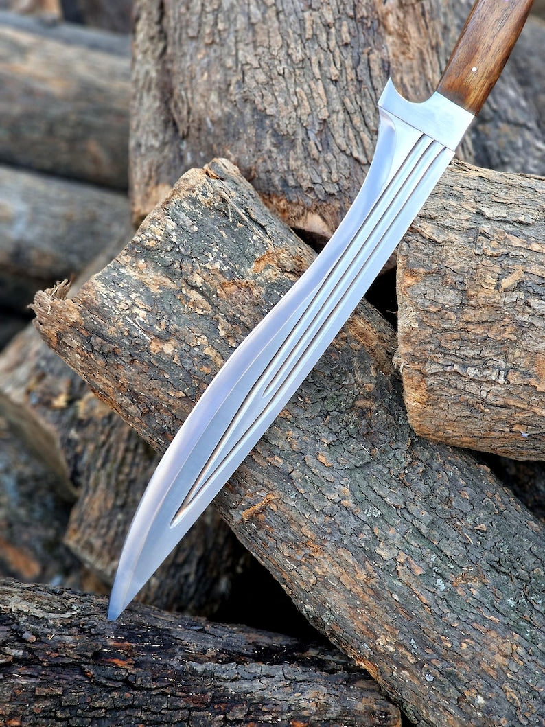 A curved silver steel knife with a wooden handle, resting on a pile of rough logs outdoors.
