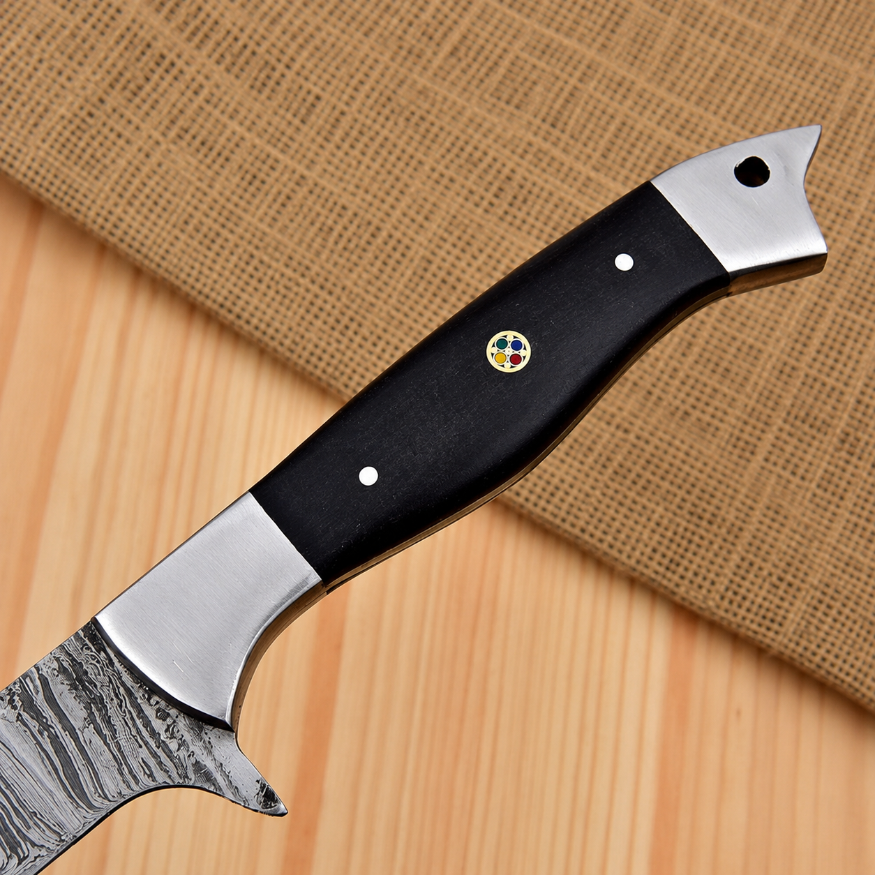 A close-up of a kitchen knife with a patterned steel blade, a black handle, and silver metal bolsters, resting on a light wood surface with a burlap cloth in the background.