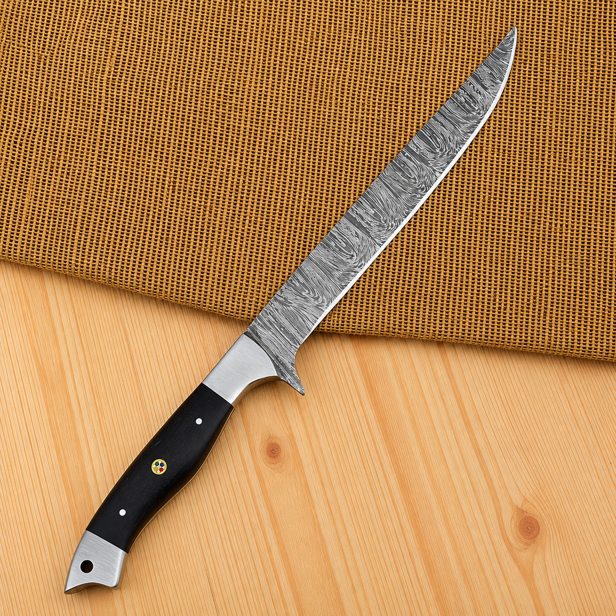 A long carving knife with a patterned Damascus steel blade and a black handle, placed diagonally on a light wood surface with a woven brown mat behind it.