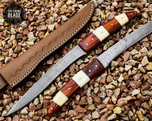 A handmade fillet knife with a Damascus steel blade and a natural wood handle, displayed on a bed of pebbles.