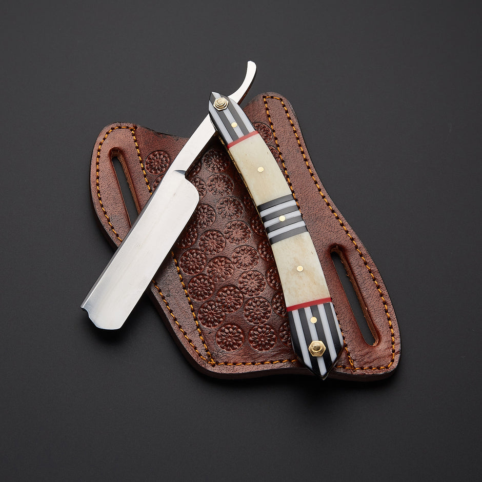 A straight razor with a polished silver blade and a decorative handle made of cream bone, black and white striped wood, and red accents, resting on a brown leather sheath with floral tooling against a dark background.